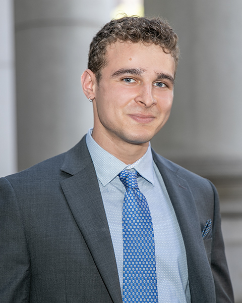 Benjamin is smiling outside, wearing a light blue shirt, blue blazer, and blue and white tie.