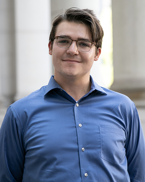 Noah is standing outside, smiling, wearing glasses and a blue button-down shirt.