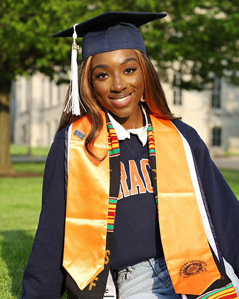 Nicole is smiling, sitting outside in the grass, wearing a graduation cap with a white tassel, a blue sweatshirt, light blue jeans, and yellow sash.