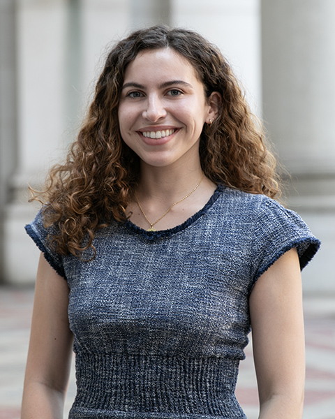 Olivia is smiling, standing outside wearing a blue top.