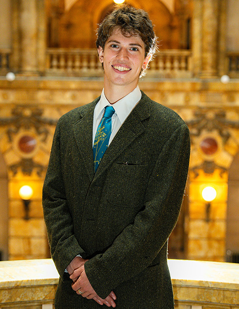 Person smiling, wearing a green blazer, white shirt and blue and yellow tie.
