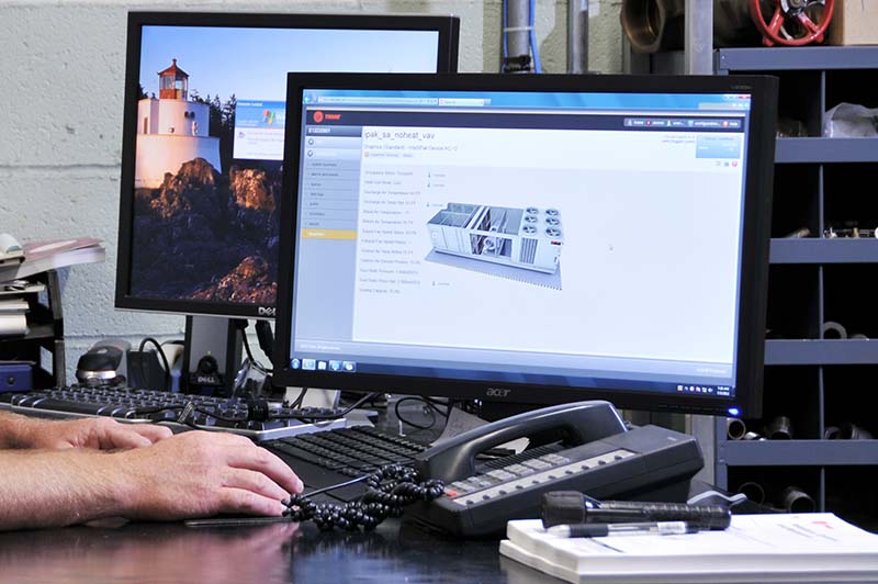 Two hands are visible in from of two computer screens, keyboards, a phone and a flashlight. Shelves with supplies are in the background. 
