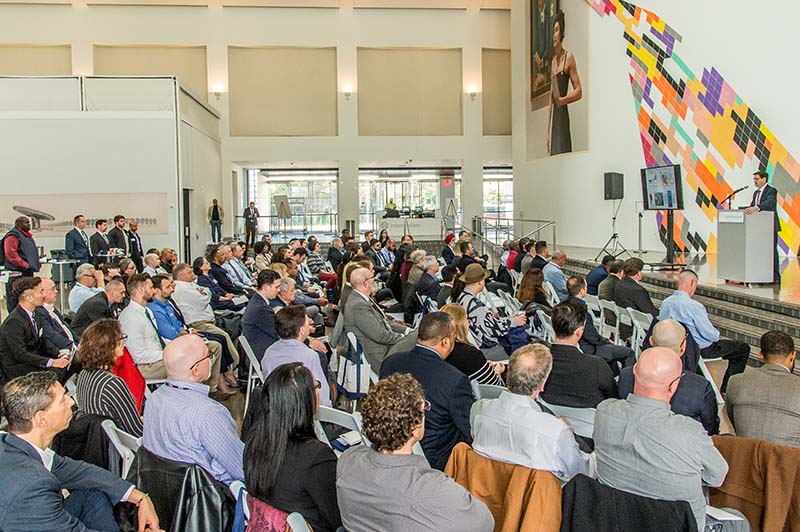 A lecture being conducted in a large open area with a stage and a diverse group of participants. On stage is the lecturer and a screen. 