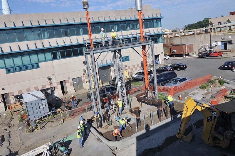 Workers in hard hats and reflective vests are digging at a site using shovels and an a small backhoe construction vehicle, as the lead contractors look on in an elevated platform. This excavation, done at the Bowery Bay wastewater treatment plant, is for a new digester gas line.