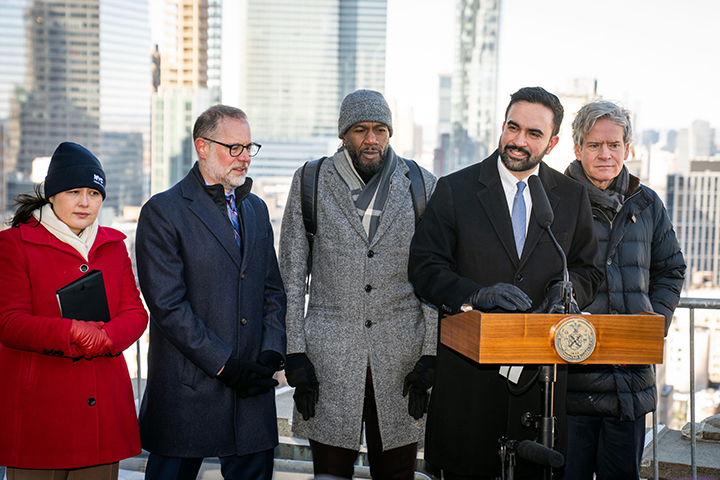 Mayor Mamdani and other city officials on the roof of the Manhattan Municipal Building with a  view of the city in the background, every one is wearing coats.
