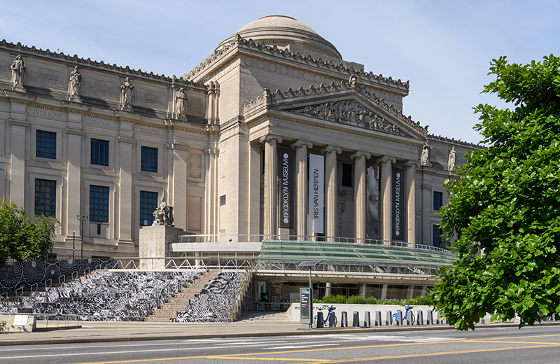 Exterior view of the Brooklyn Museum, showing its classical stone fa&ccedil;ade, columns, dome, entrance banners, front plaza, and a leafy tree along the street in the foreground.