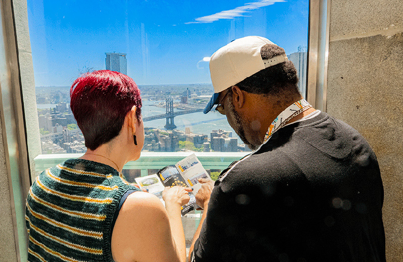 Two visitors look out from a high window while reviewing a pamphlet, with the Manhattan Bridge and East River visible in the distance.
