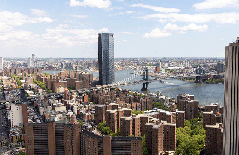 A panoramic view of Lower Manhattan and Brooklyn, featuring the Manhattan Bridge spanning the East River under a partly cloudy sky.