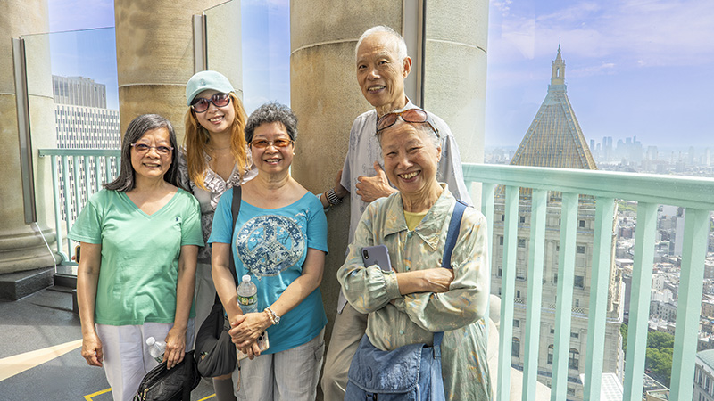 Five smiling visitors pose on the Centre 360 observation deck, with the Manhattan Municipal Building cupola and city skyline visible behind them.
