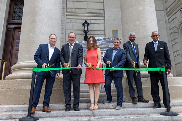 City officials including five men in suits and a woman in a red dress all holding scissors to cute the green ribbon in front of them on the steps of court building.