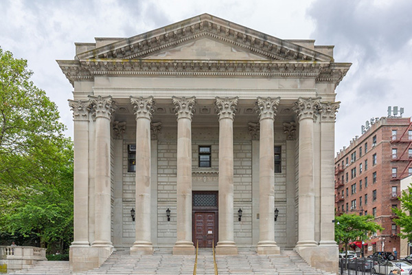 The front of a French Renaissance style, temple-fronted limestone courthouse with multiple pillars on a street with trees and residential buildings.
