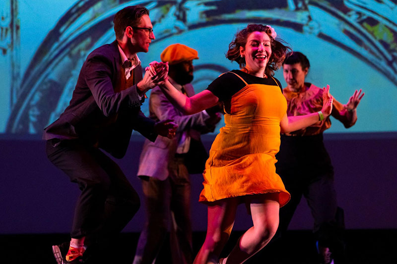 A performer wearing a yellow dress is on stage. Three others are in the back. 
