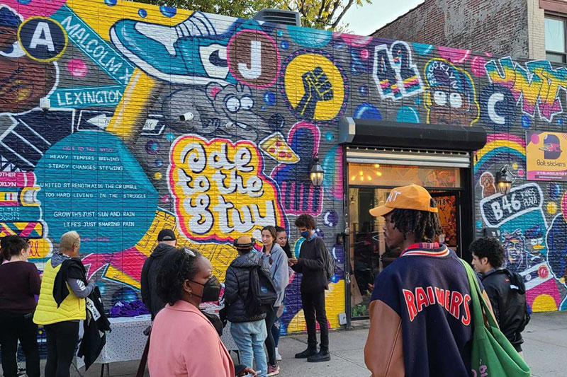 People gather in front of a colorful mural celebrating Bed-Stuy and its icons