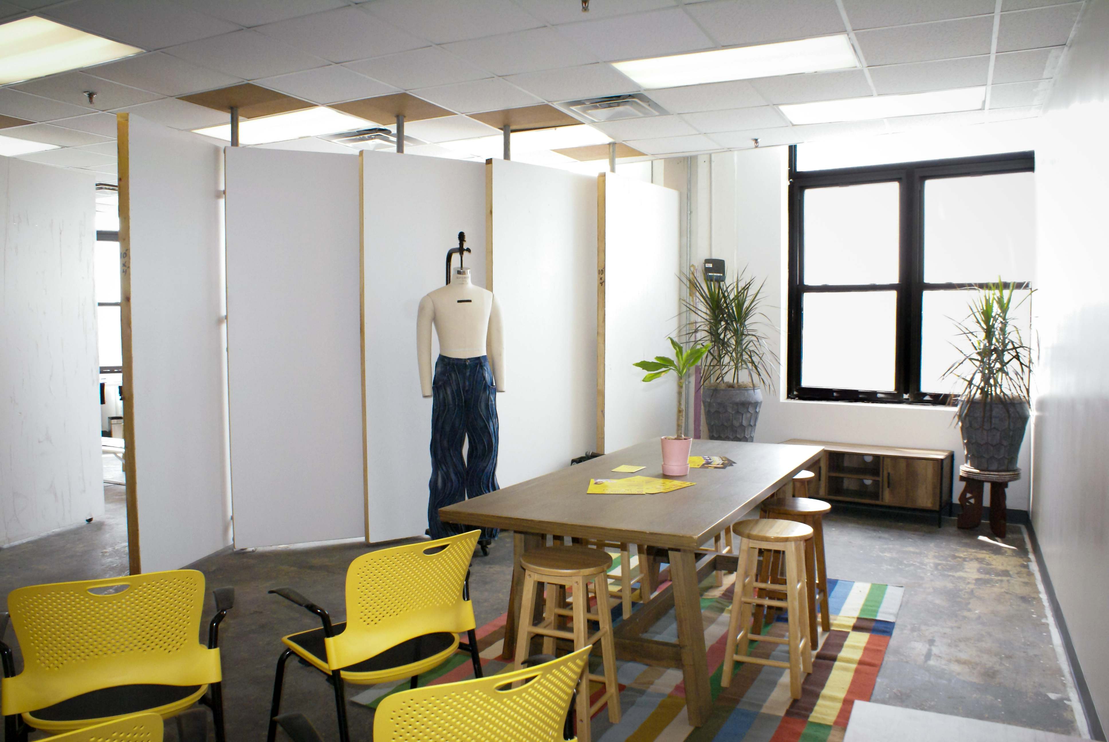 A studio space with a cured wall, dress form, table, plants, and yellow chairs.