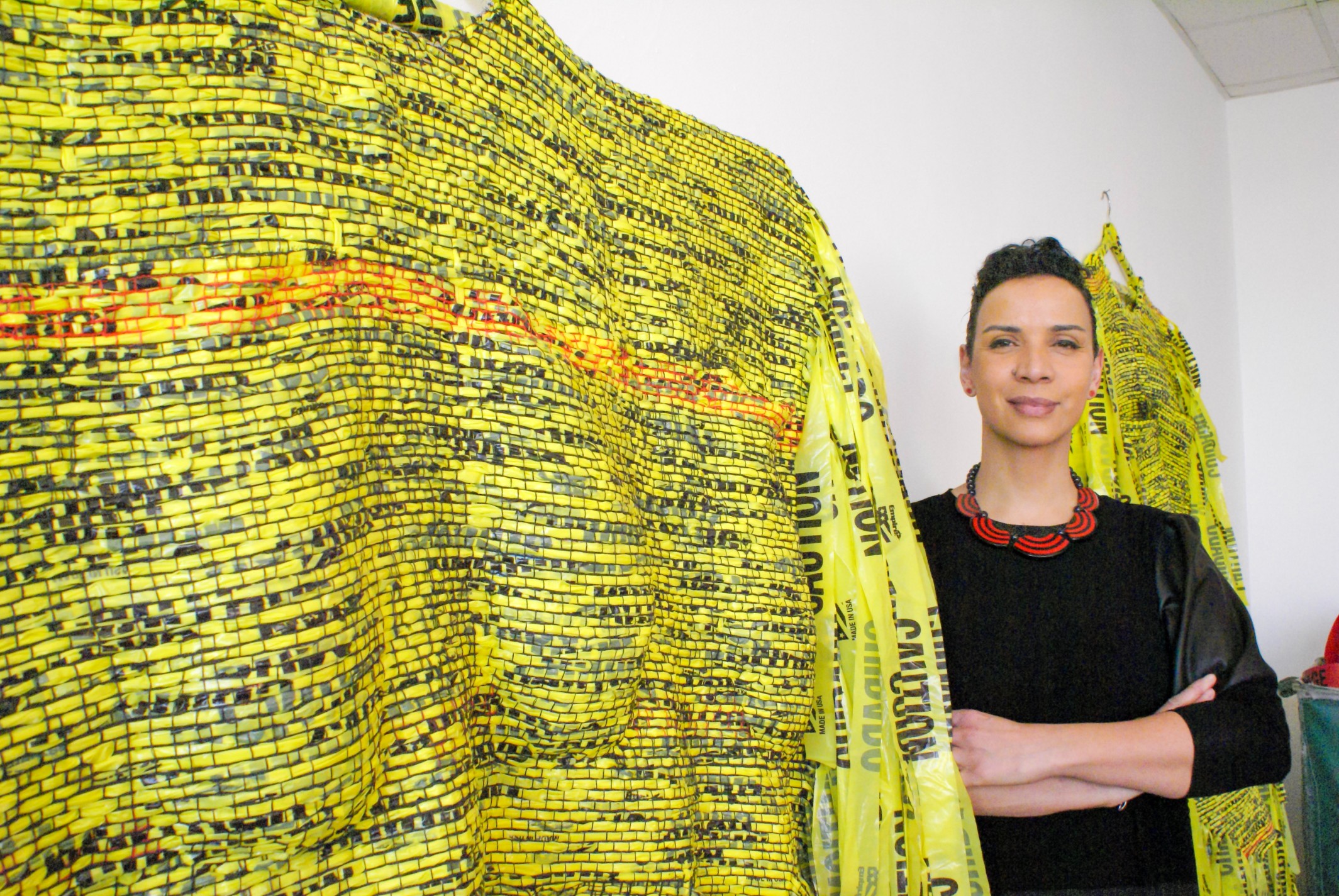 Borinquen Gallo stands beside a yellow woven piece made from caution tape. 