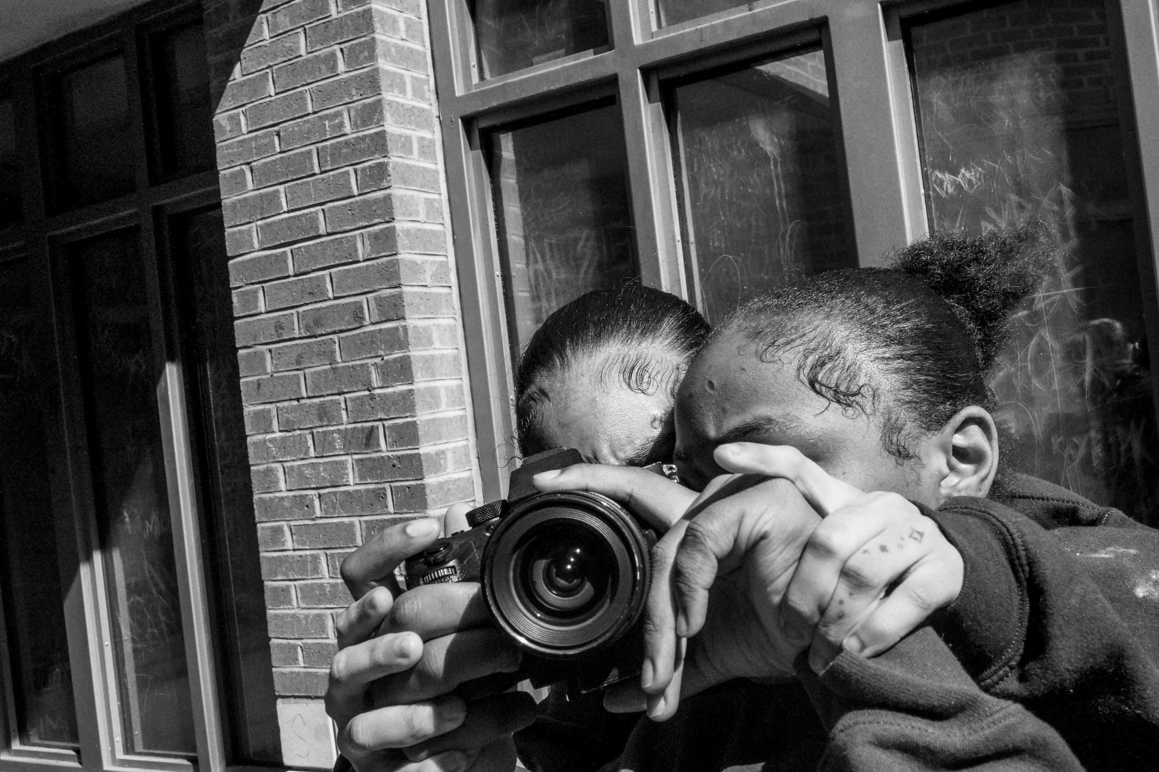 A black and white photo of two teens looking through one camera together.