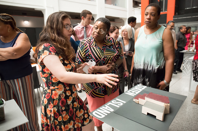 A student points at a model on a table while two DDC staff members look on.