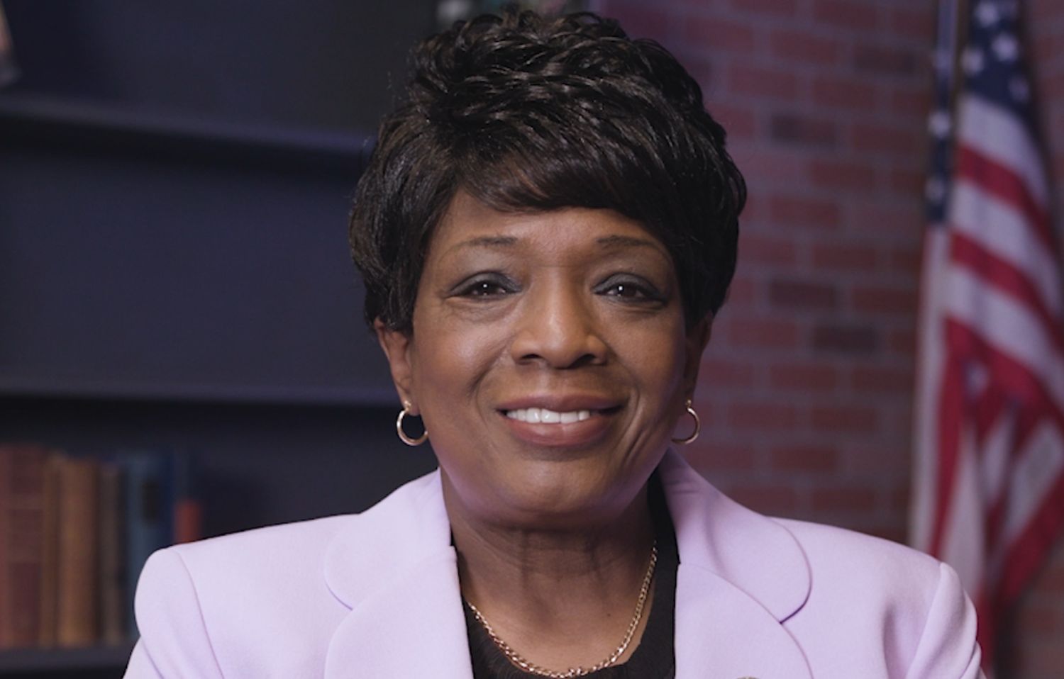 Black woman in lavender blazer smiling in front of blurred U.S. flag backdrop.
                                           