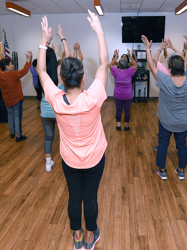 A group of older adults taking a fitness class stretch their hands up to the ceiling.