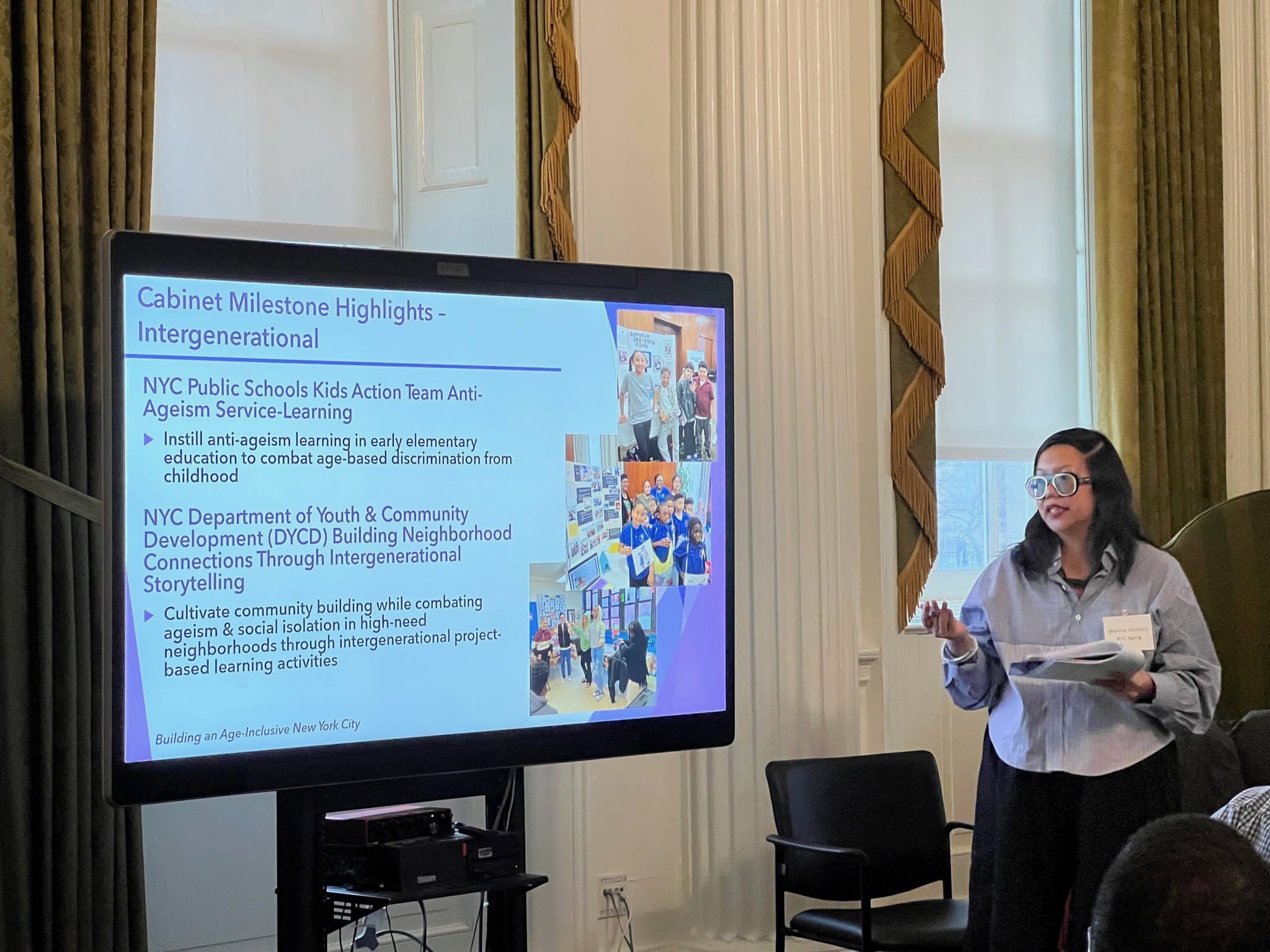 A woman presenter stands beside a screen displaying a slide on intergenerational programs from NYC Public Schools and DYCD during a Cabinet meeting.