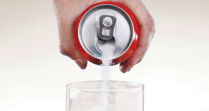 Photograph of a hand holdinga soda can pouring sugar into a drinking glass.