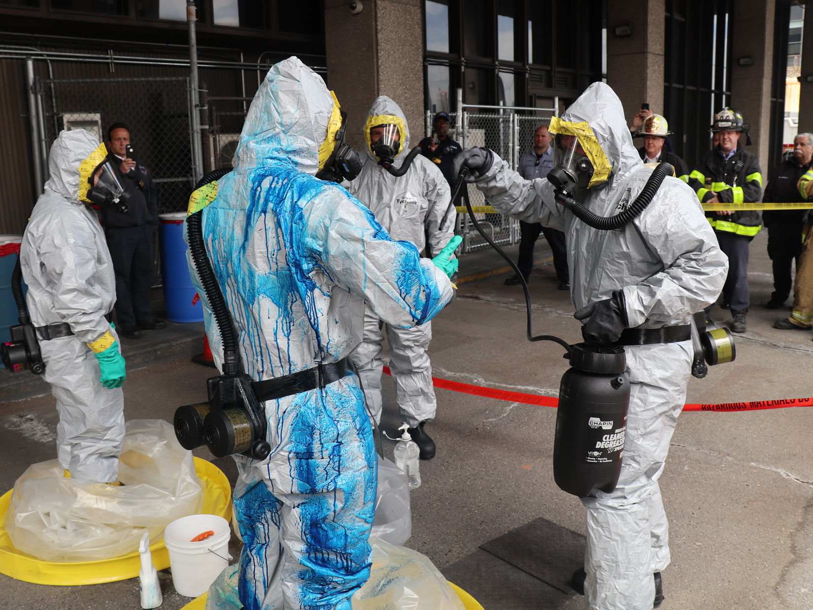emergency responders dressed in haz-mat suits, with one covered in bleach, are spraying each other with liquids