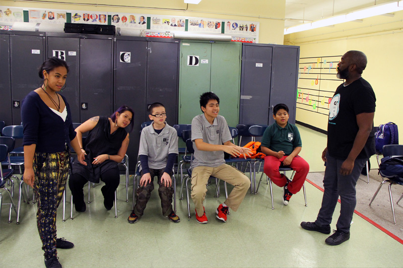 four students sit down in chairs while one stands and looks ahead in classroom