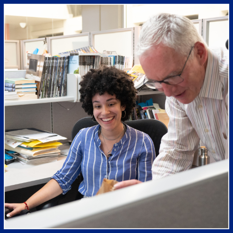 A female participant wearing a striped light-blue shirt sits on a computer while speaking to a male instructor. The Summer Youth Employment Program.