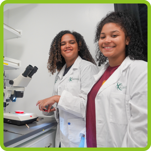 Two young women in lab coats stand in front of a microscope