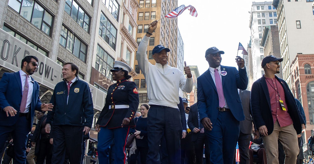 Mayor Adams and Commissioner Hendon lead a past Veterans Parade holding small American flags.