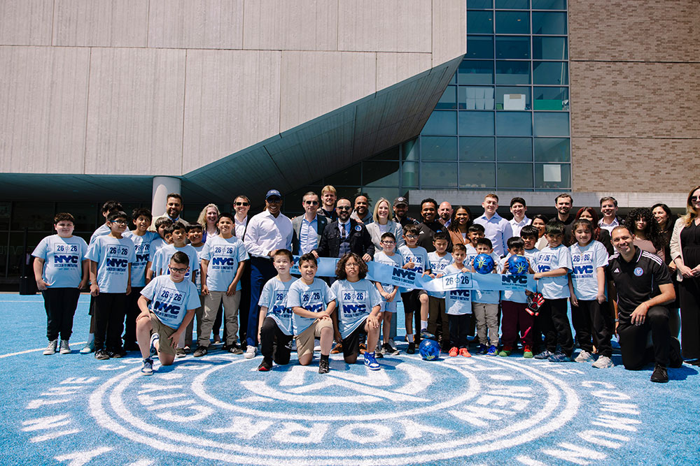Mayor Eric Adams, supporters of the new soccer pitch, and students pose for a photo with a blue cut ribbon for the opening of the field.