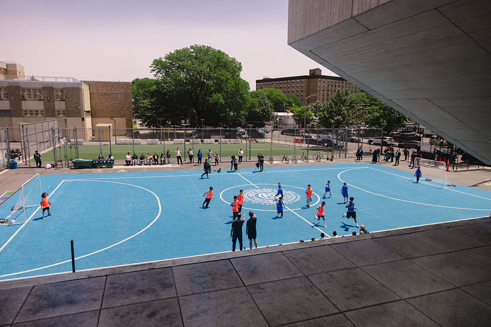 An overhead photo of a blue turf field being used by young students in a game of soccer.