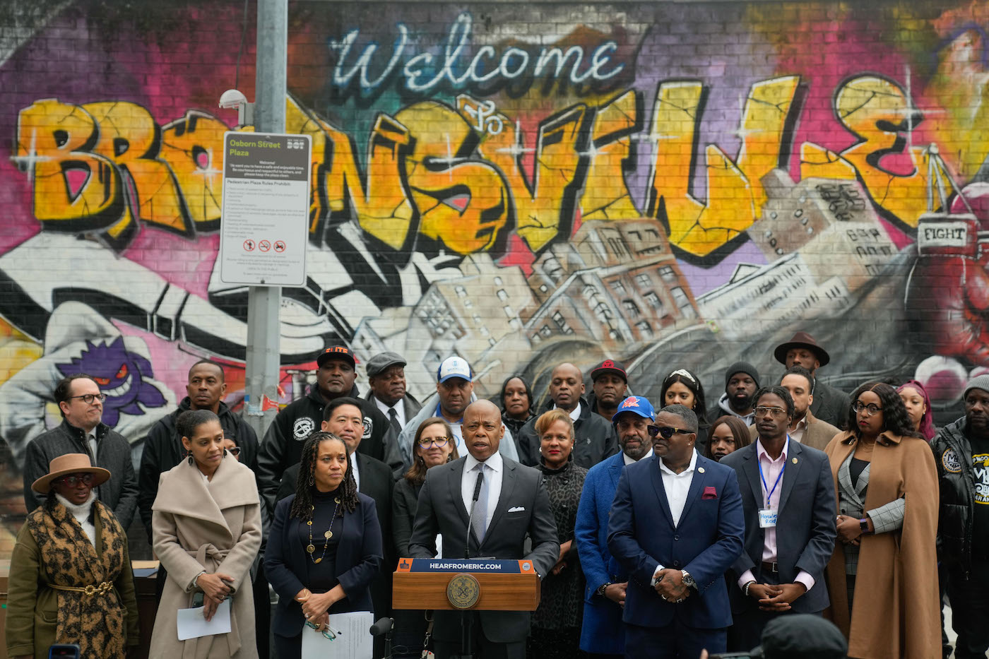 Mayor Adams delivers remarks from a podium in front of a mural and is surrounded by adminsitration officials