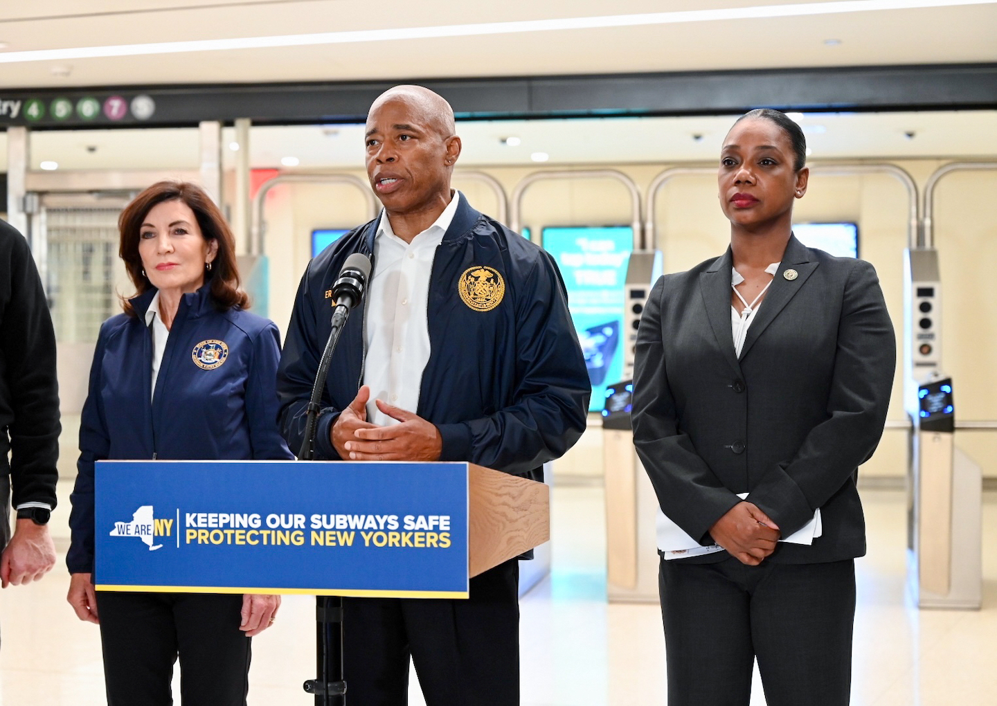 Mayor Adams standing next to two women, speaking from a podium in the subway.