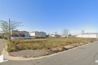 A grassy, vacant lot surrounded by roadways, with homes visible in the background.