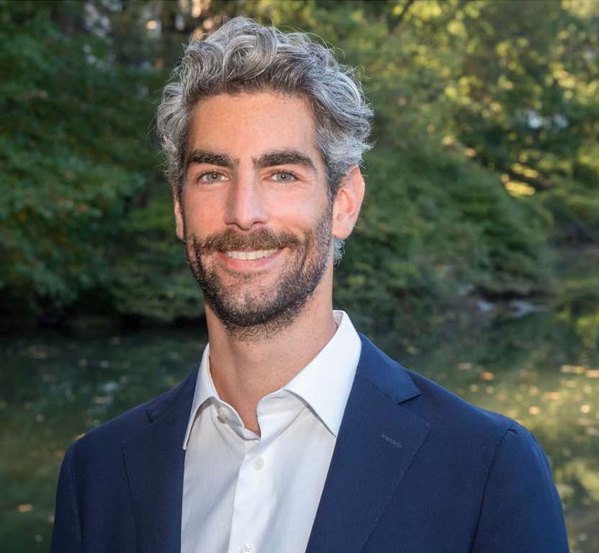 Headshot of Adam Phillips, First Deputy Commissioner, smiling with curly gray hair and beard in a suit stands outdoors against a lush green, sunlit background
