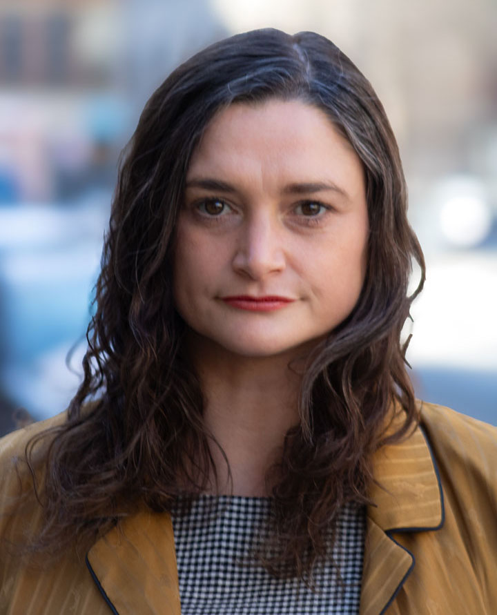 Portrait of Celeste Hornbach, Executive Advisor for Special Projects with long brown hair, wearing a tan jacket over a checkered top. She is standing in front of a blurred urban background.