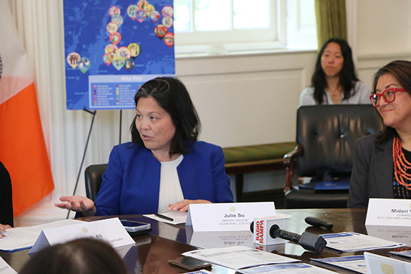 A woman wearing a blue shirt is sitting at a round table with others, speaking and pointing to the person on her right. Another woman is in the background listening to the speakers. Behind the woman are an NYC flag and a poster board with artwork.