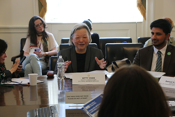 A woman is sitting at a round table alongside others and speaking to audience members. A man sits to her left at the table. Audience members are sitting behind the round table listening to speakers. There are flyers on the table