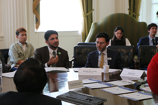 A man in a black blazer sits at a round table with others and speaks to audience members. A man sits to his left at the table. Audience members are sitting behind the round table listening to speakers. There are flyers on the table.