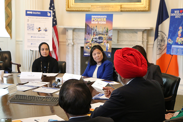 A group of people sits at a round table and speaks among themselves. Behind them are poster boards, an NYC flag, and an American flag.