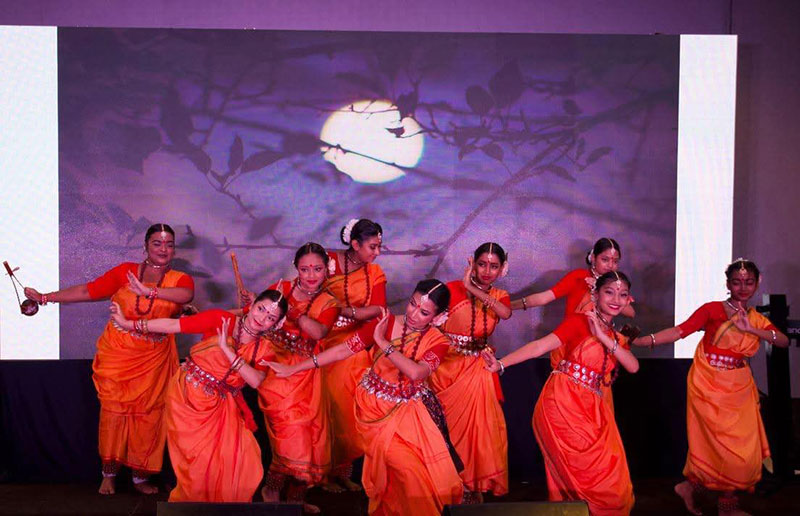 Several women dressed in traditional Bengali clothes dance on stage with a screen in the background.