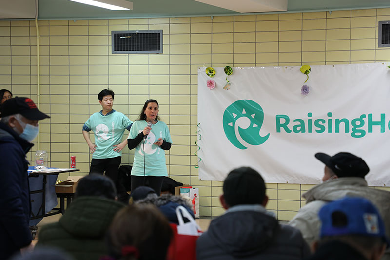 Inside a room with people sitting and listening to a speaker at the front of the room make an announcement. A woman stands with a man and makes an announcement