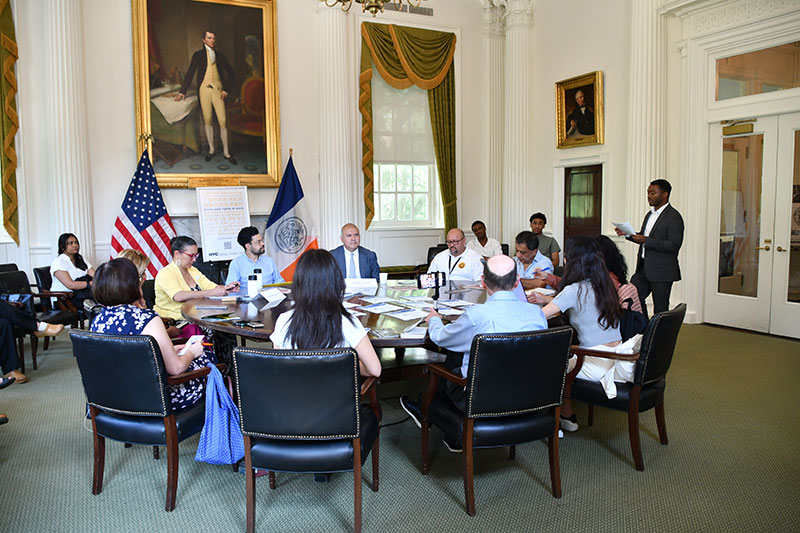 A group of city workers and local reporters sit around a large round table inside a large room during an event while listening to a speaker. A man standing on the side is looking at the paper in his hand