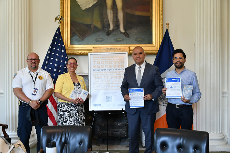 Four people stand in front of a sign about workers' rights in multiple languages and two flags while holding up resources