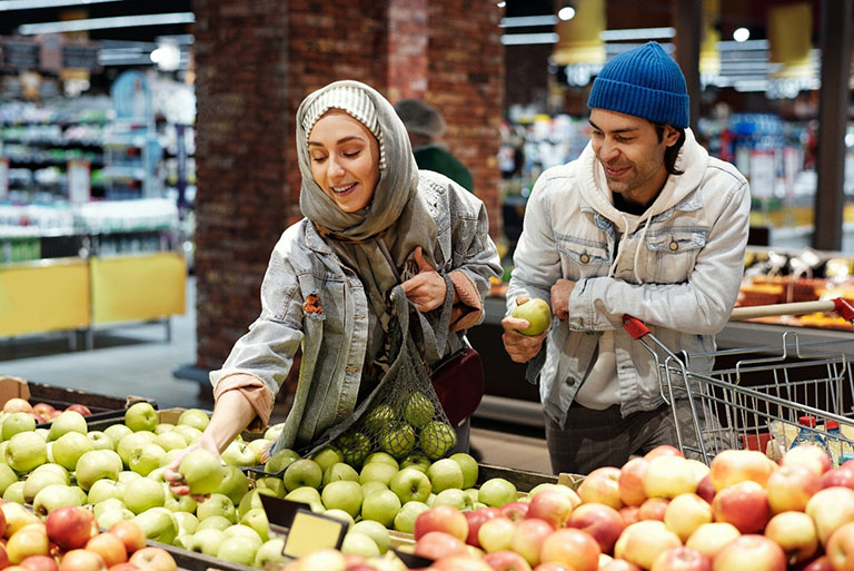 Two people at a supermarket. The woman is picking up an apple while the man is standing next to her with the cart and holding an apple