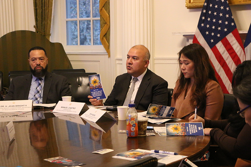 Three people are sitting at a table in front of a flag. A man is speaking while others are listening.