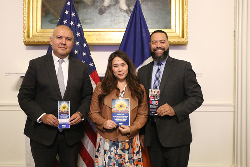 Three  people standing together holding up a brochure and standing in front of two flags