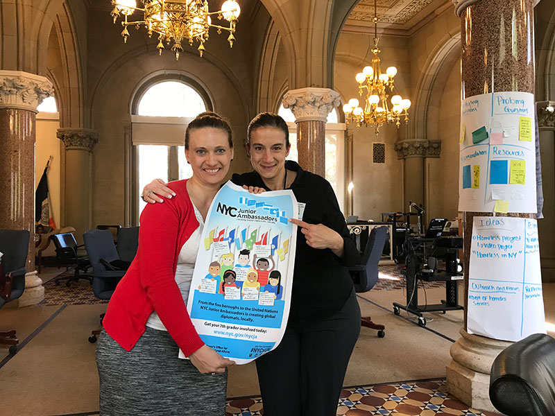 Two teachers posing with a NYC Junior Ambassadors poster