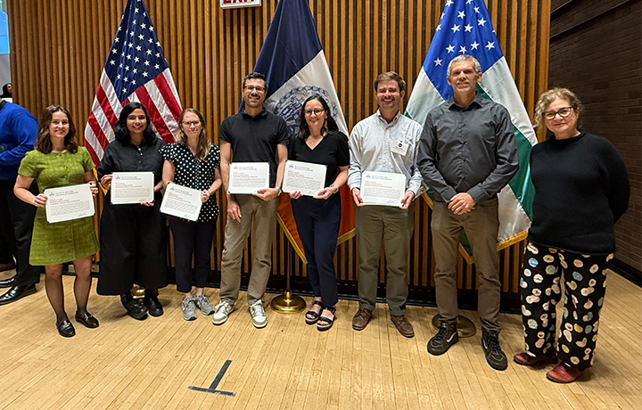 Group of eight smiling people, some holding certificates, with wood paneling and three flags in the background